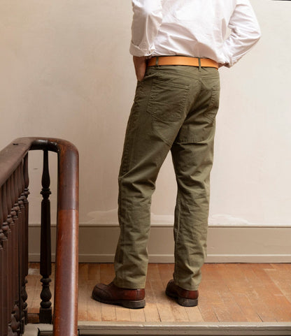 A person wearing Best Made Co.’s Field Pant Special Edition Olive Drab stands with their back to the camera on a wooden floor near a staircase, paired with a white shirt, tan belt, and brown shoes.