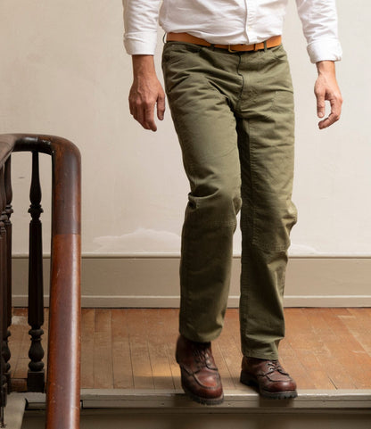 Someone wearing Best Made Co. Field Pant Special Edition Olive Drab, a white shirt, and brown shoes walks down a wooden staircase with a wooden handrail, only their lower half visible.