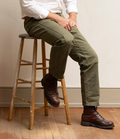 A person in a white shirt, Field Pant Special Edition Olive Drab by Best Made Co., and brown boots sits on a wooden stool against a light wall and wood floor. Their face is not visible.