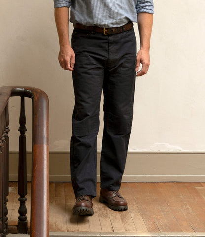 A man stands indoors on a wooden floor near a staircase, wearing a light blue striped shirt and Best Made Co. Field Pant Special Edition Navy, paired with brown shoes. His arms are relaxed at his sides, and his face is not visible.
