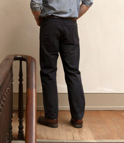 A person stands indoors with their back to the camera, wearing a striped shirt and Best Made Co. Field Pant Special Edition Navy, next to a wooden railing on a worn hardwood floor.