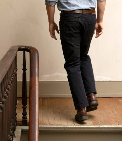 A man in a blue striped shirt, brown belt, and Best Made Co. Field Pant Special Edition Navy walks upstairs on a wooden staircase with a brown handrail, seen from behind.