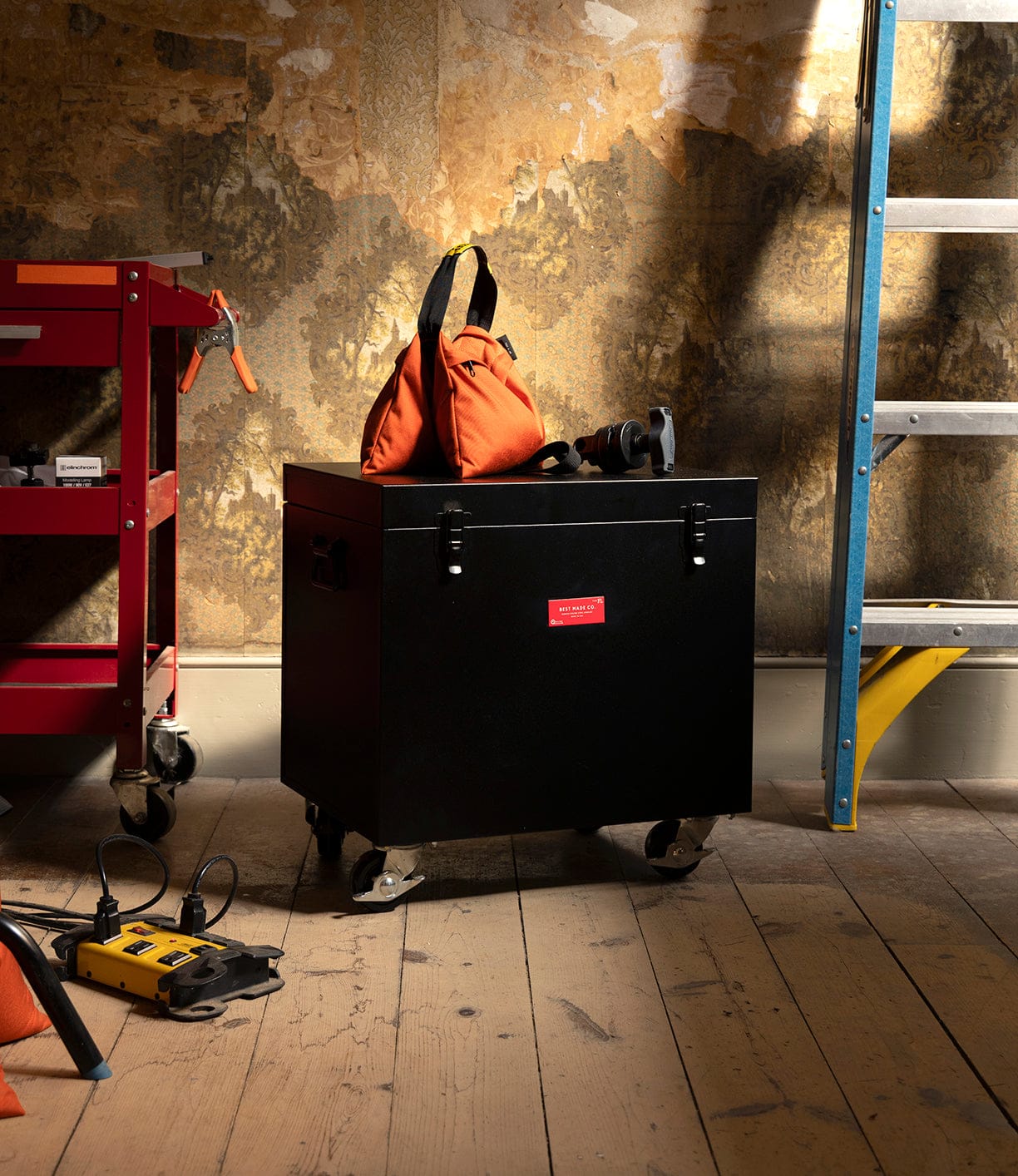 A black Best Made Co Rolling Footlocker with a red label sits on a wooden floor, topped with an orange bag, tape measure, and flashlight. Nearby are a red cart, yellow ladder, and power tool, all set against a peeling wall.