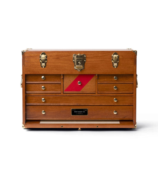 A man in glasses and a cap leans on a Best Made Co. Gerstner 41D Founder’s Edition machinist chest, hand on head. Tools and frames decorate the wall, while two axes lie on the table in front. Black and white photo.