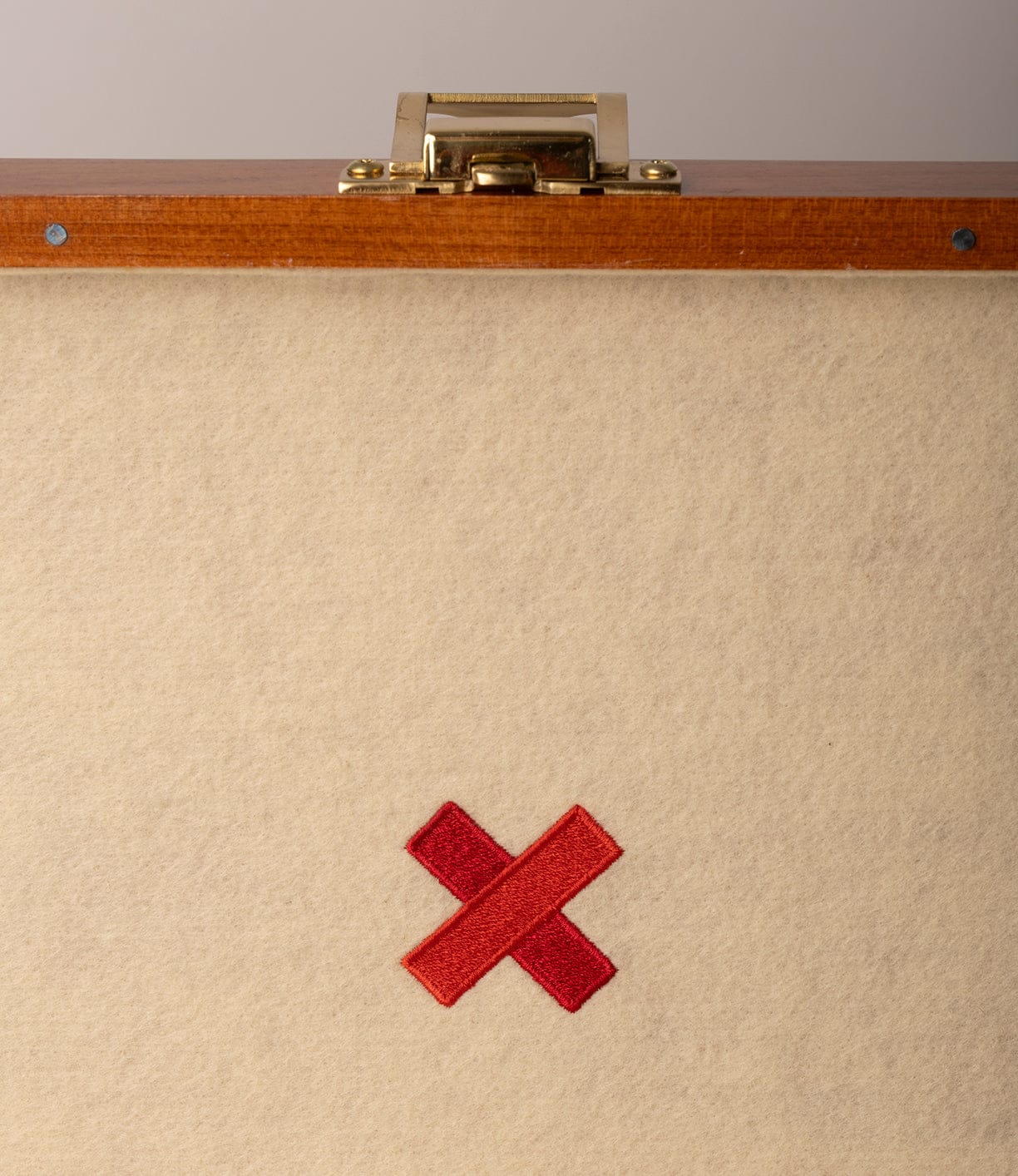 A beige surface with a red embroidered X near the bottom sits beneath the wooden edge and brass latch of a closed Best Made Co. Gerstner 41D Founder’s Edition chest at the top of the image.