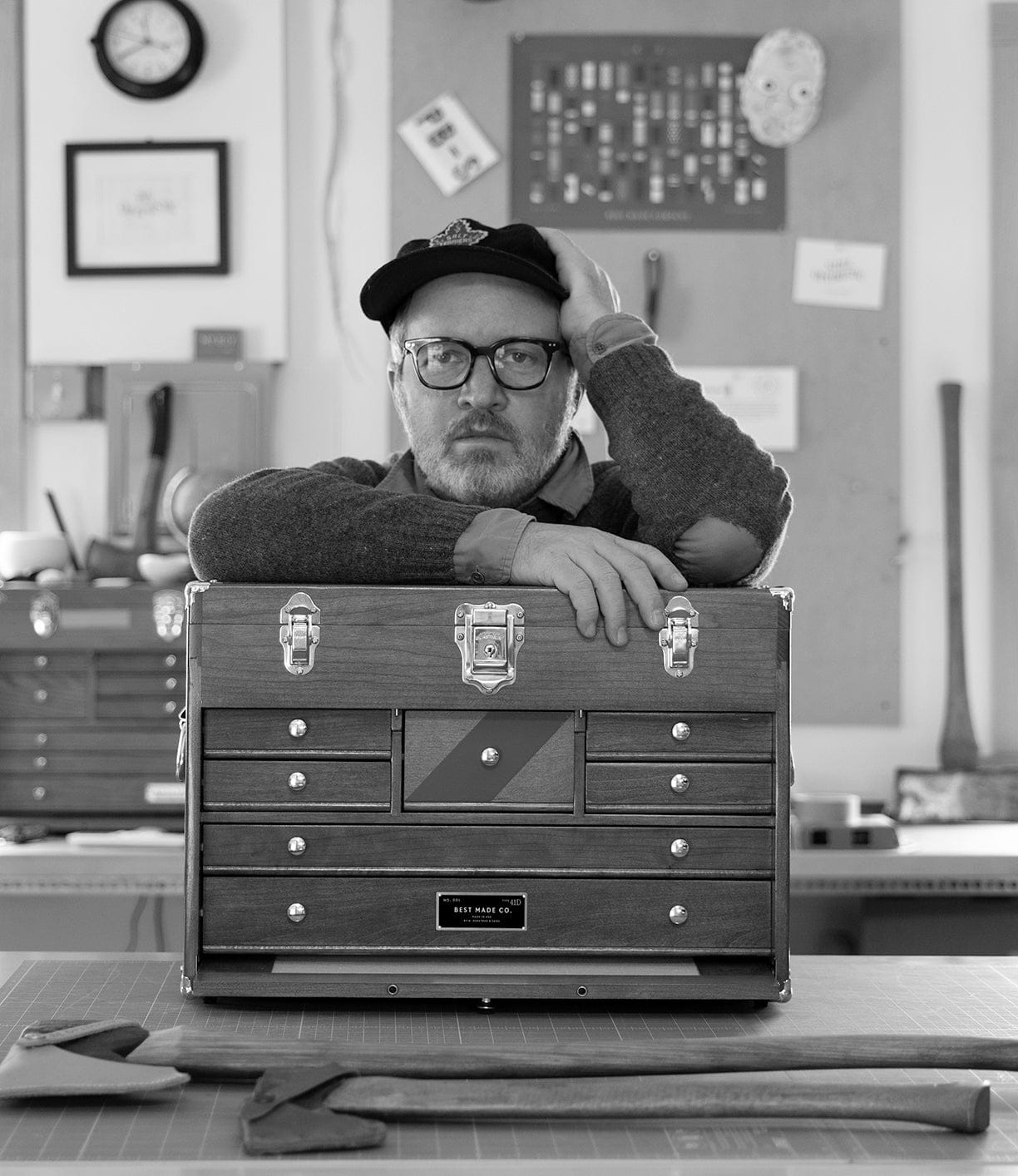 A man in glasses and a cap leans on a Best Made Co. Gerstner 41D Founder’s Edition machinist chest, hand on head. Tools and frames decorate the wall, while two axes lie on the table in front. Black and white photo.