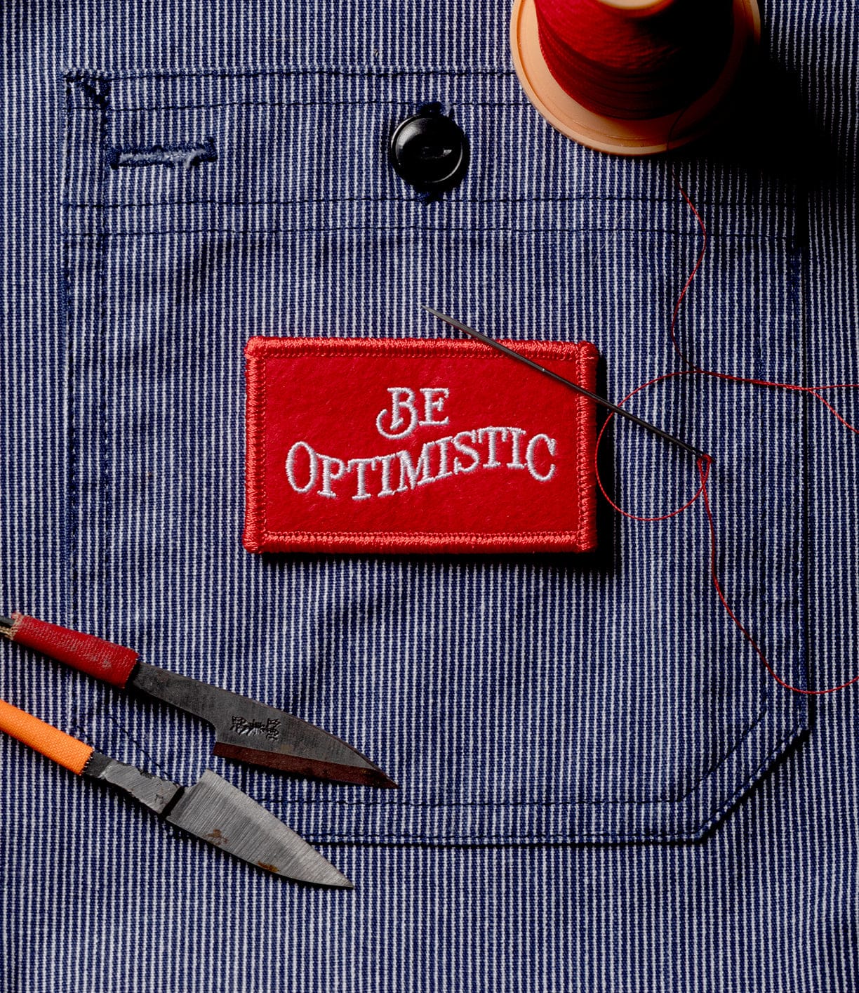 The Best Made Co. Be Optimistic Patch, featuring bold white letters on red, sits atop a blue and white striped pocket beside a needle with red thread and survival tool-inspired sewing gear, capturing the spirit of outdoor adventure.