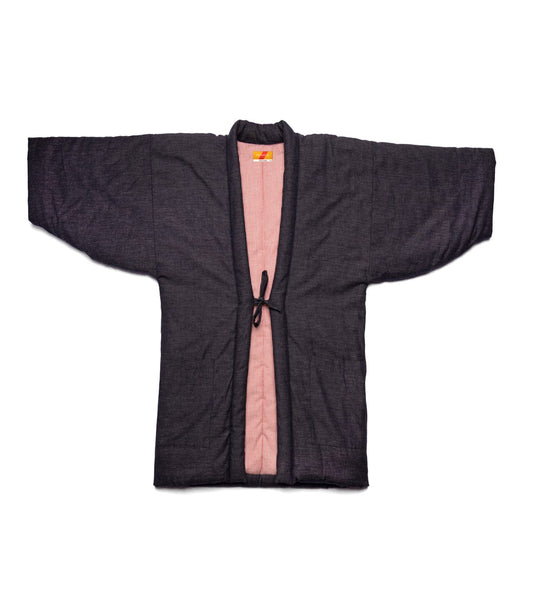 A man in a blue shirt, olive green pants, and the Best Made Co. Quilted Hanten Jacket with red-and-white lining stands with one hand in his pocket, looking to the side against a textured backdrop.