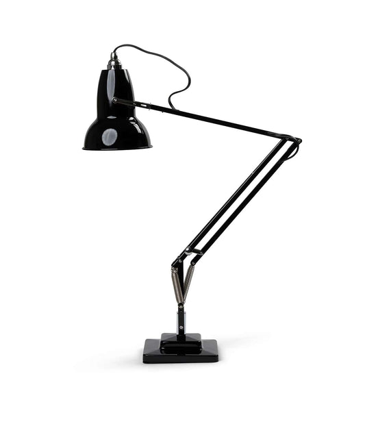 The Best Made Co Anglepoise Original 1227 Desk Lamp in black sits on a metal shelf with a red notebook, black glasses, a mug of pens, and a STILL OPTIMISTIC white box—all against a textured wall for classic British task lamp style.