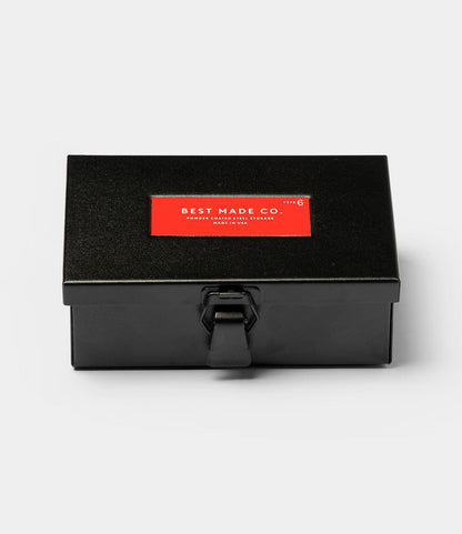 The Best Made Co. Type 6 Strongbox in matte black, a rectangular steel box with a latch and red lid label, offers durable storage and is shown against a plain white background.