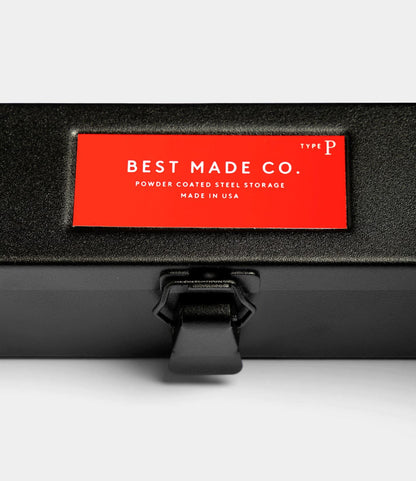 A close-up of the Type P Strongbox - Matte Black by Best Made Co., a durable powder-coated steel box with a latch and red label—perfect for lasting pencil storage and organization.