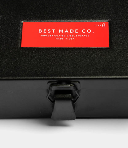 A close-up of the Type 6 Strongbox - Matte Black by Best Made Co., a rugged black steel box with a latch and red “BEST MADE CO. Powder Coated Steel Storage Made in USA Type 6” label—ideal for durable storage and organization.