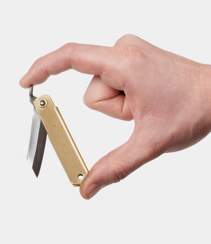 A hand holds an open Mid Brass Higo Knife by Best Made Co., with "BEST" engraved on the handle, set against a plain white background.