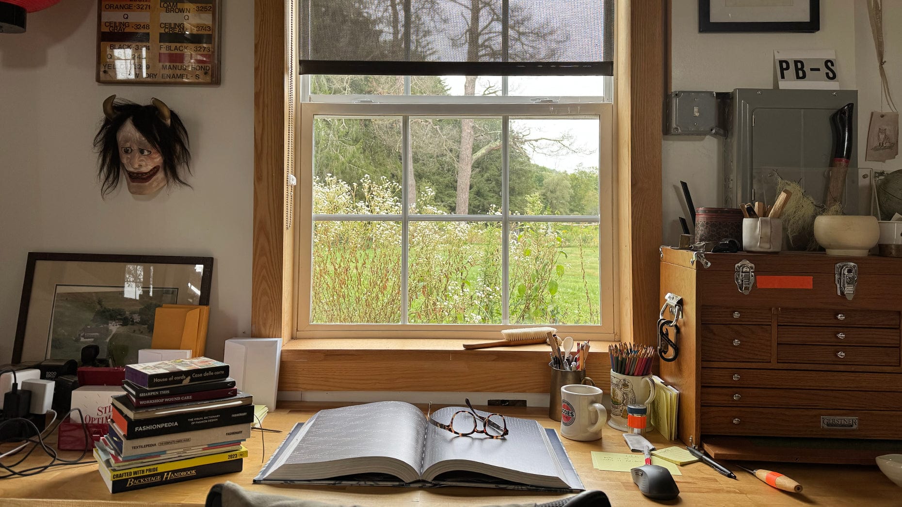 A wooden desk with an open book, glasses, mugs, pens, and stacked books sits by a window overlooking a green garden. A tribal mask, art supplies, photos, and a wooden chest decorate the cozy workspace.
