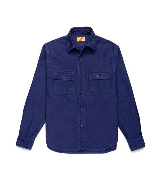 A man with light brown hair and a mustache stands against a textured wall, wearing the Best Made Co Indigo Moleskin Work Shirt under a blue jacket and olive green pants, with his hands behind his back.