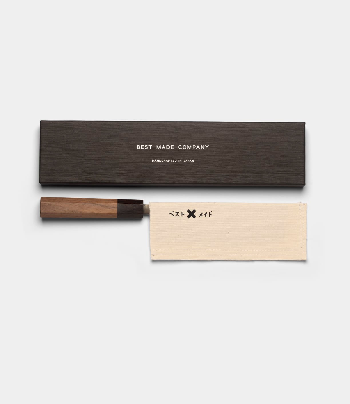 The Aogami Nakiri Knife by Best Made Co., featuring a wooden handle, rests beside a black Best Made Co. box and a beige cloth adorned with black symbols. Expertly crafted from Blue Paper Steel No. 2, this knife merges traditional Japanese design with outstanding cutting performance.