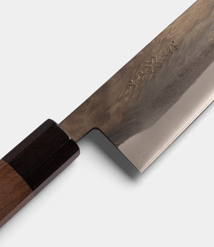 A close-up of the Best Made Co. Aogami Santoku Knife, showcasing its dark wooden handle and lustrous Blue Paper Steel blade adorned with engraved markings near the handle, set against a light background.