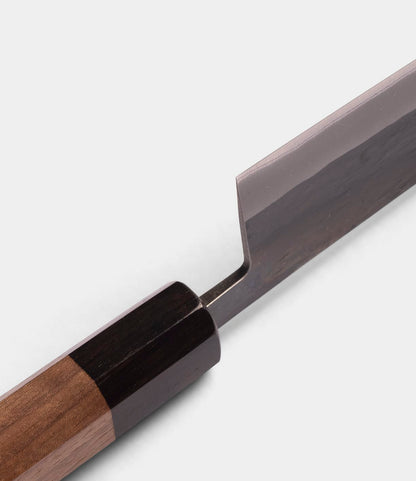 Close-up of the Best Made Co. Aogami Santoku Knife, featuring a wooden handle with a dark bolster and part of its shiny, straight-edged Blue Paper Steel blade, set against a plain light background.