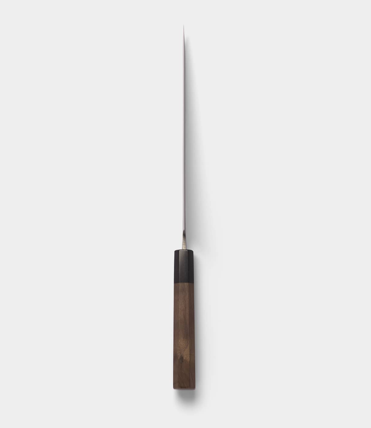 The Best Made Co. Aogami Gyuto Knife is displayed from above, highlighting its slender silver blade—made with Blue Paper Steel No. 2—and its rectangular wooden handle, which casts a narrow shadow on a simple light gray background.