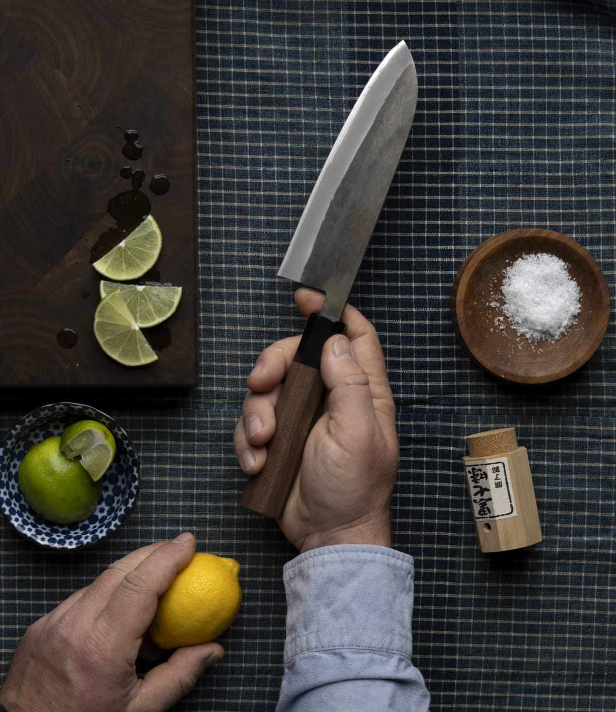 A person holds the Best Made Co. Aogami Santoku knife, featuring a Shou Sugi Ban handle, in one hand and a lemon in the other. Sliced limes, a bowl of salt, and a wooden spice container rest on a checkered cloth.