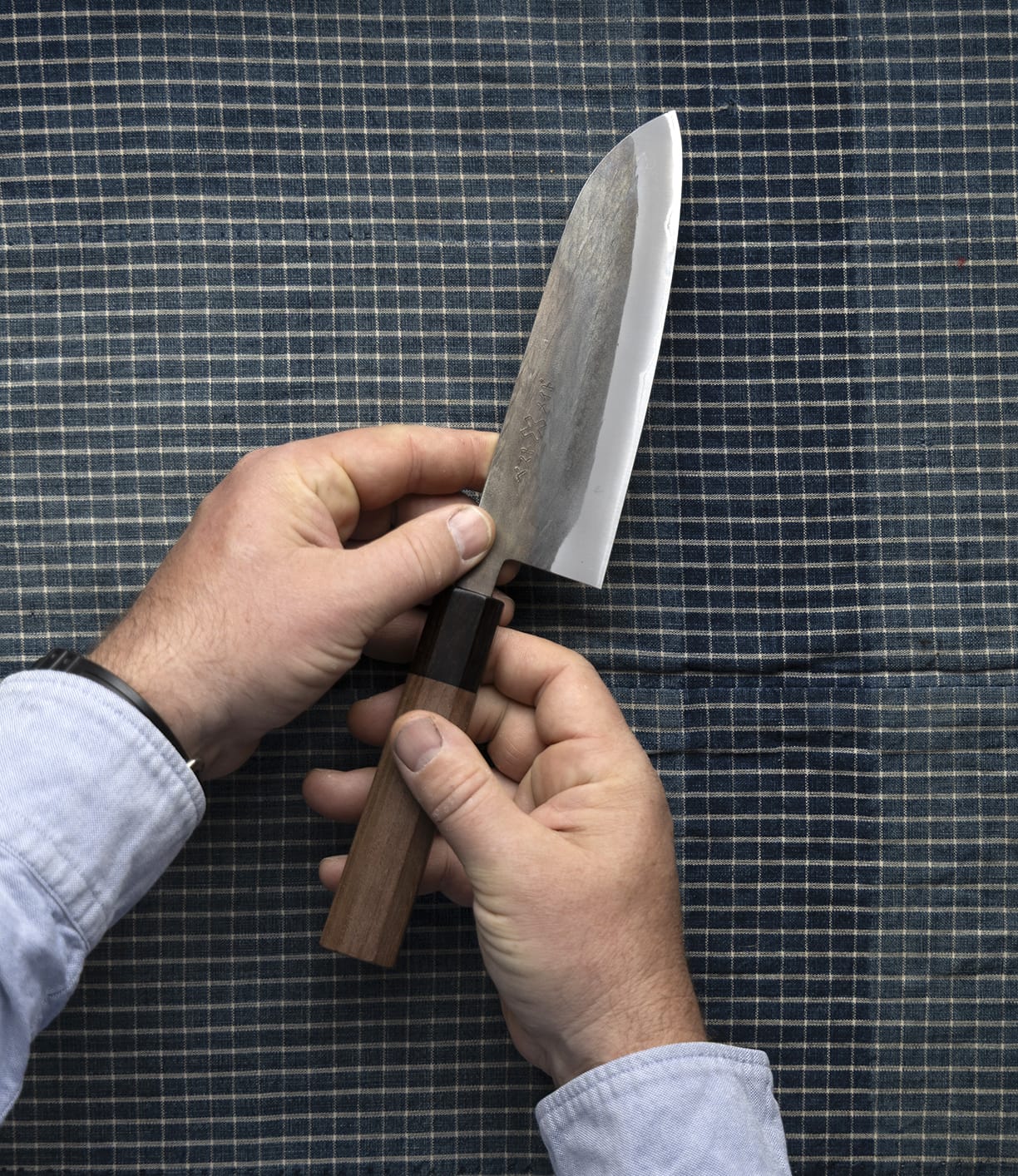 A person holds the Best Made Co. Aogami Santoku Japanese kitchen knife, featuring a wooden handle, against a blue and white checkered fabric background.