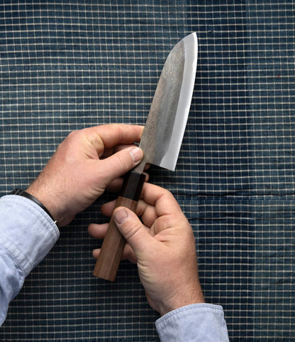 A person holds the Best Made Co. Aogami Santoku Japanese kitchen knife, featuring a wooden handle, against a blue and white checkered fabric background.