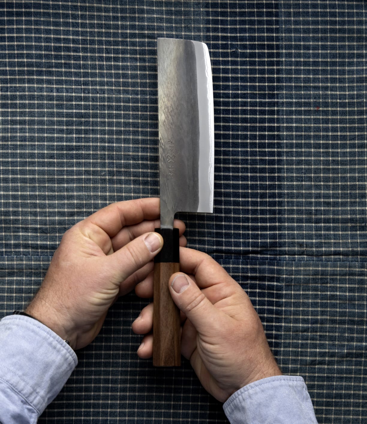 Two hands hold the Best Made Co. Aogami Nakiri Knife—featuring a wooden handle and a Blue Paper Steel No. 2 blade—over a blue and white checkered fabric background.