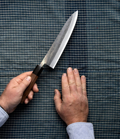 A person holds a sharp Best Made Co. Aogami Gyuto knife in one hand while their other hand rests flat on a blue checkered cloth surface, preparing to cut or chop.