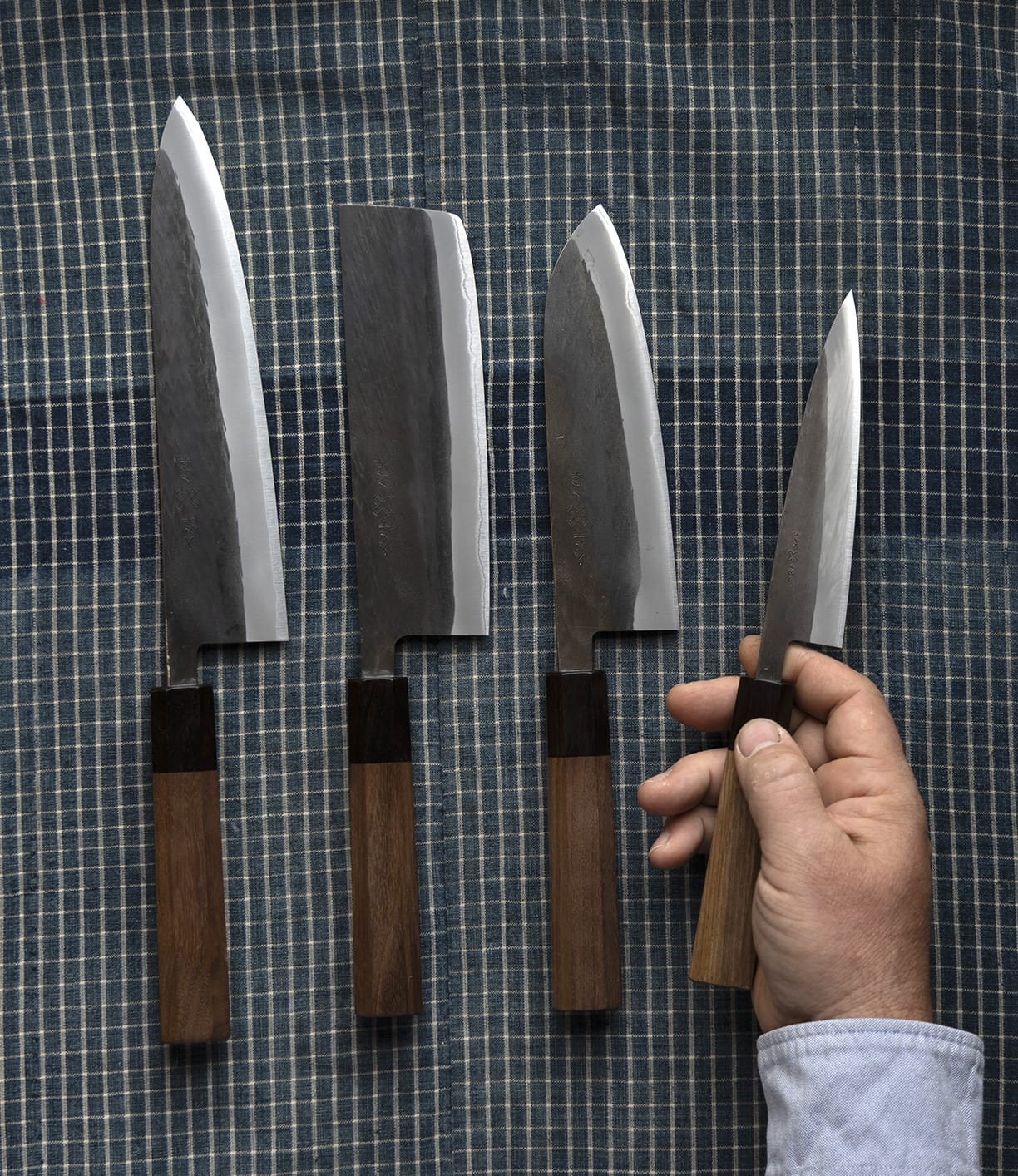 A hand holds the Best Made Co. Aogami Gyuto Knife, distinguished by its wooden handle, next to three more of the same knives arranged on a blue checkered cloth. Crafted with Blue Paper Steel No. 2 for exceptional sharpness and precision, these knives are perfect for any culinary task.
