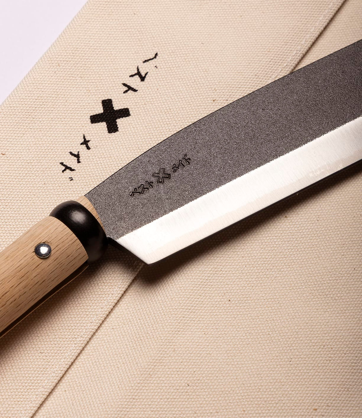 A close-up of the Best Made Co. Sayanata Hatchet, a Japanese-style kitchen hatchet with a versatile blade and a wooden handle, resting on a beige cloth featuring black printed characters and symbols.