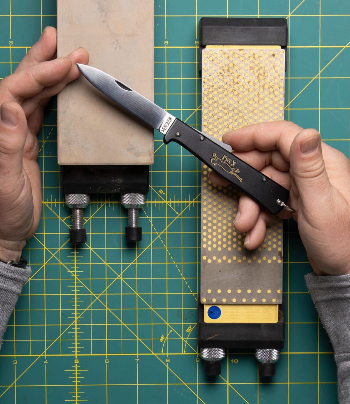 A person sharpens a Best Made Co. K55K Mercator carbon steel knife with a sharpening stone in one hand and another stone beside it, both on a green cutting mat with a yellow grid.