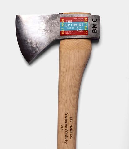 A hand-forged axe by Best Made Co., The Optimist, features a wooden handle and an American 5160 carbon steel head with a colorful label: THE OPTIMIST HUDSON BAY AXE.
