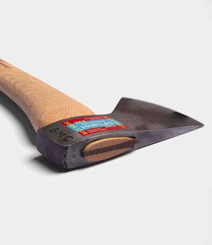 A close-up of The Optimist by Best Made Co., a Hudson Bay axe with a wooden handle, durable 5160 carbon steel head, and colorful sticker label on top, shown against a plain white background.