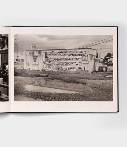 Open Range by Best Made Co.: a black-and-white photo of a building in Alpine, Texas, its mural mirrored in a puddle beneath a cloudy sky—capturing the quiet rural mood reminiscent of Robert Adams’ photography.