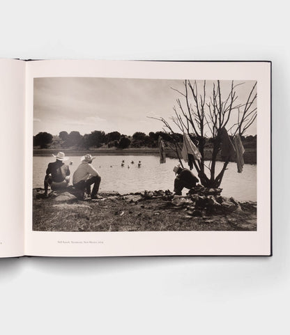 The Best Made Co. "Open Range" photography book lies open, showing a black-and-white image of three people—two with cowboy hats—by a lake, clothes on a bare tree, wooden boats nearby, and swimmers in the background.