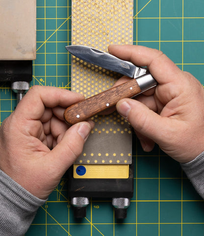 A person sharpens a Best Made Co. Otter-Messer Pocket Knife with a sharpening stone on a green cutting mat, both hands visible holding the knife and stone.