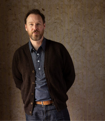 A man wears a black Best Made Co. Black Sheep Shetland wool V-neck cardigan over a grey button-down shirt and blue jeans.