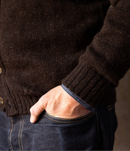Close-up of a person wearing a Black Sheep Cardigan in black Shetland wool with a crew neck and ribbed cuffs, with their hand tucked into the pocket of dark denim jeans.