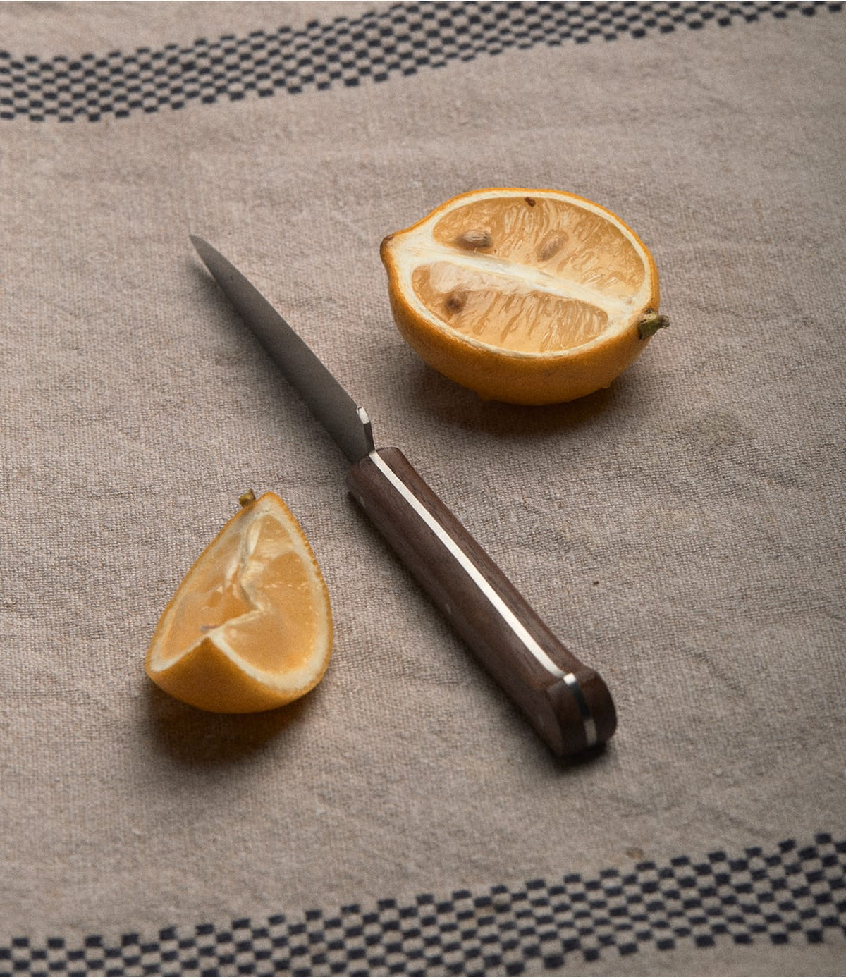 A Le P’tit Tradi paring knife with a walnut handle lying on a linen cloth next to a halved lemon and a lemon wedge.