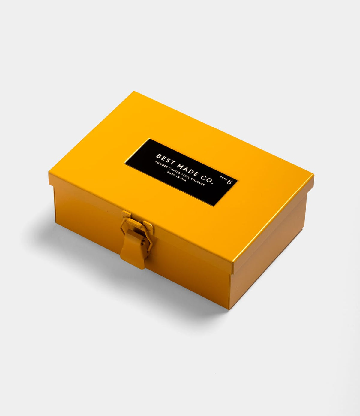 A yellow Type 6 Strongbox Yellow by Best Made Co., featuring a clasp and black rectangular label, is placed on a white background.