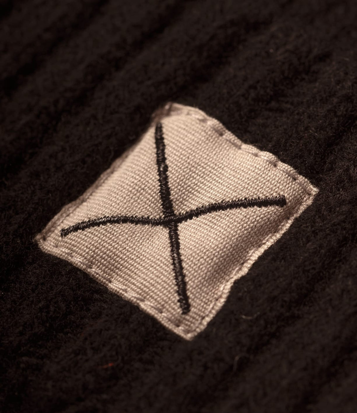 A small, square, light-colored patch with an embroidered black X on the black ribbed knit fabric of a Best Made Co. Cap of Courage.