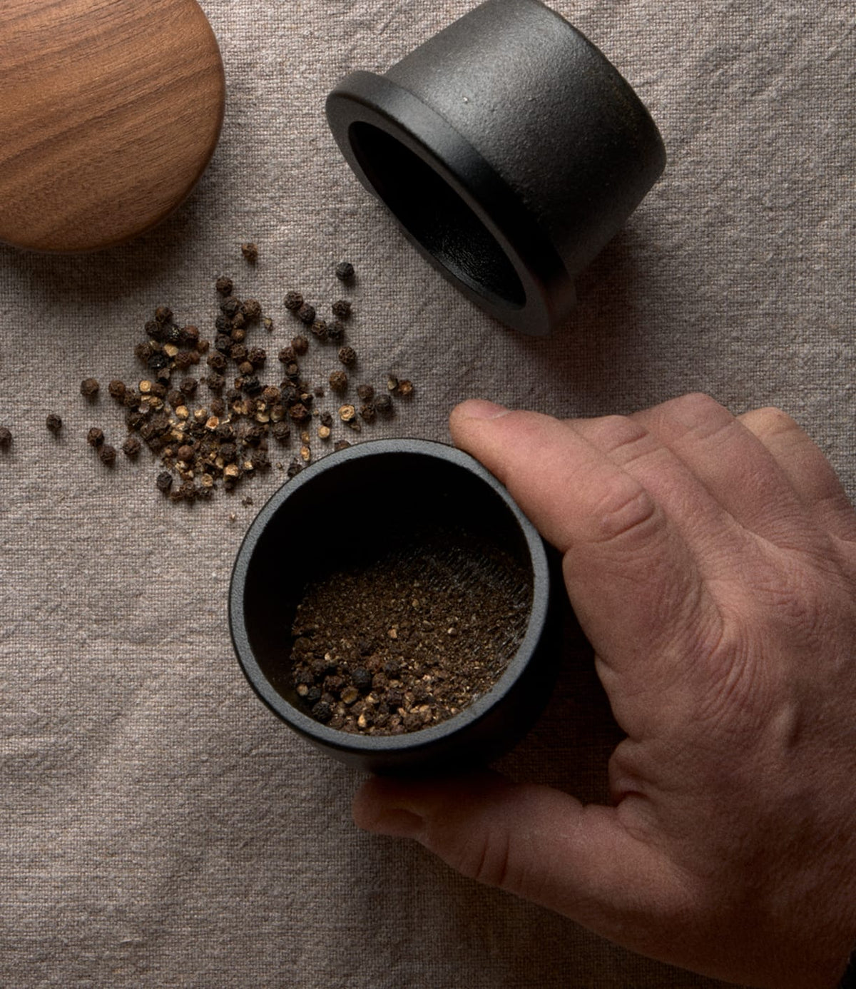 A hand holds a Best Made Co. Cast Iron Grinder filled with cracked peppercorns, next to its matching walnut lid and a pile of whole peppercorns on a linen surface.