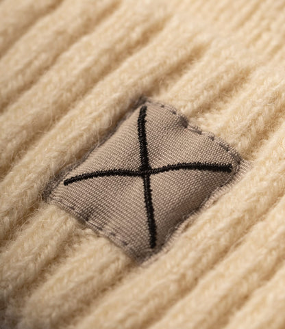 Close-up of a ribbed, off-white Shetland Cap of Courage in undyed Shetland wool, featuring a square patch embroidered with a black X.