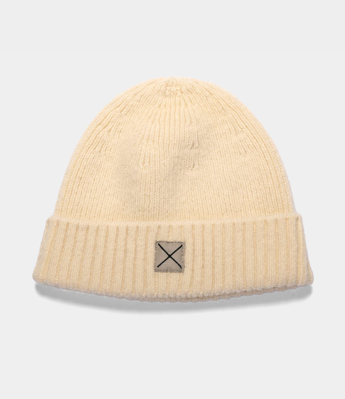 Cream-colored Shetland Cap of Courage ribbed knit beanie made of undyed Shetland wool, featuring a small square patch with an X stitch on the cuff.