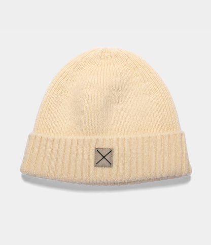 Cream-colored Shetland Cap of Courage ribbed knit beanie made of undyed Shetland wool, featuring a small square patch with an X stitch on the cuff.