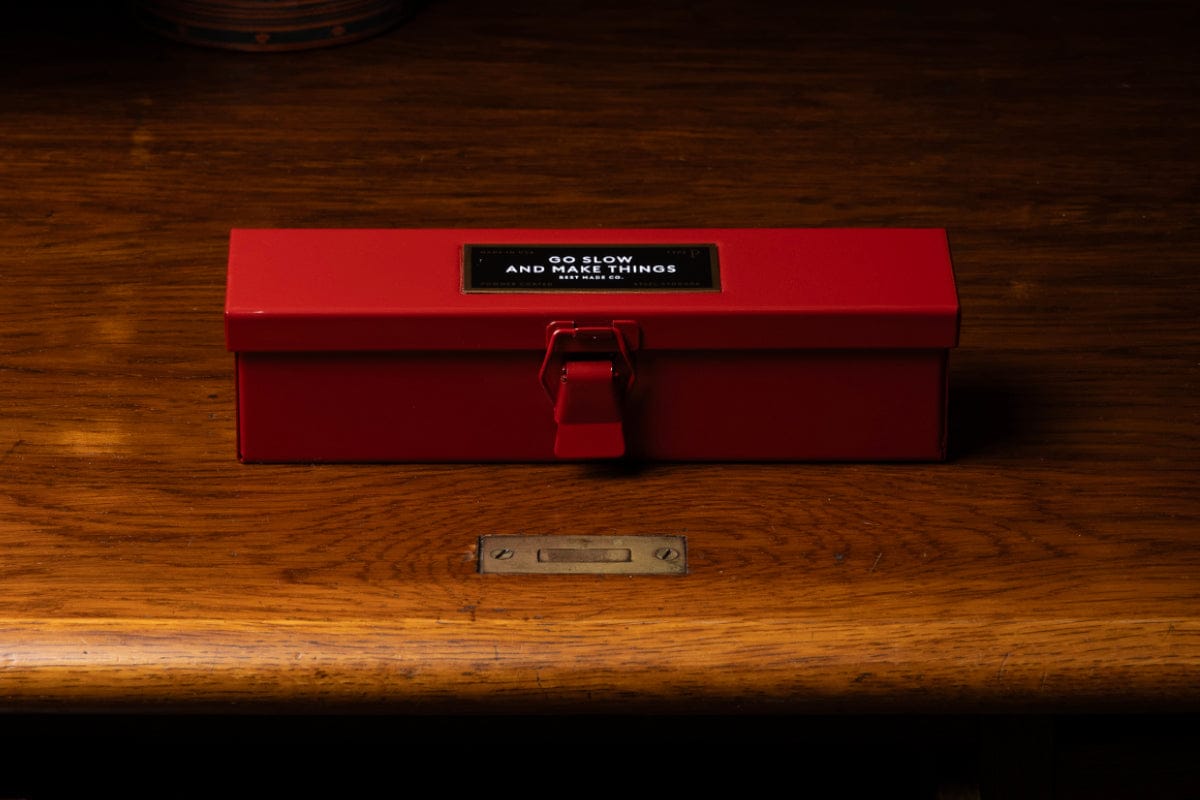 A red metal box with a latch lies on a wooden surface. The box has a black label that reads GO SLOW AND MAKE THINGS. The wood grain and a small brass nameplate are visible on the surface.