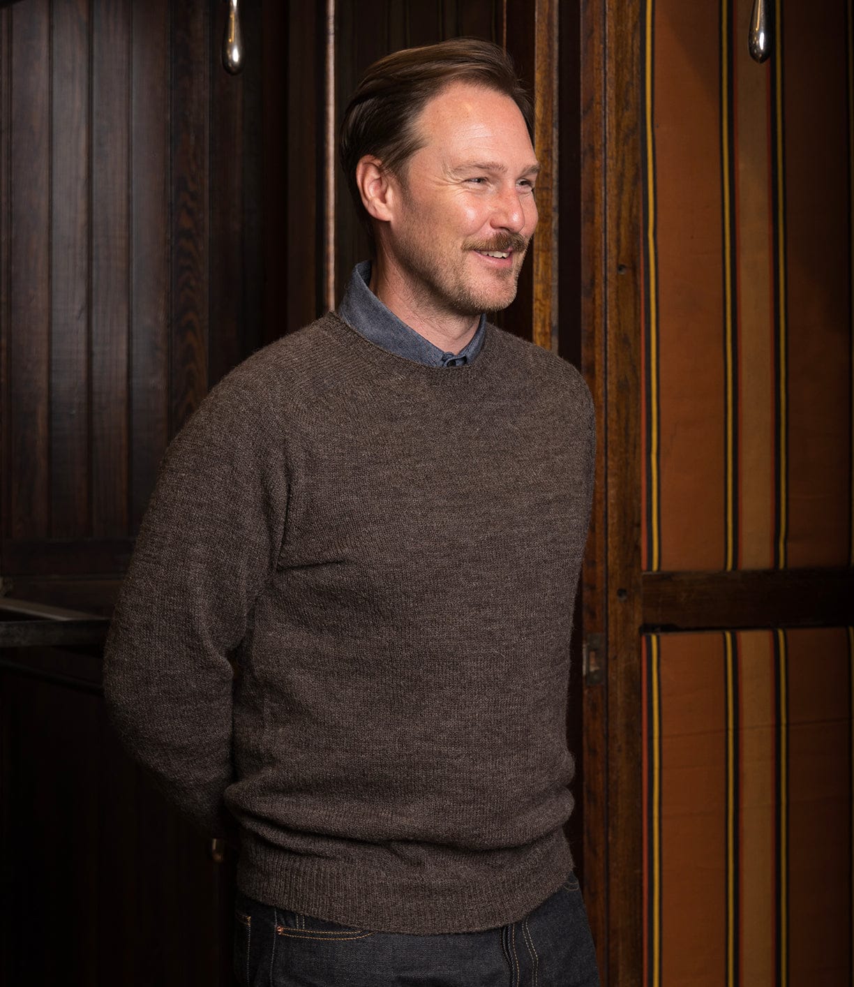 A man with light brown hair and a mustache, wearing the 5-Gauge Shetland Sweater by Best Made Co. over a blue shirt, stands indoors smiling in front of dark wooden panels and an orange-striped wall, hands behind his back.