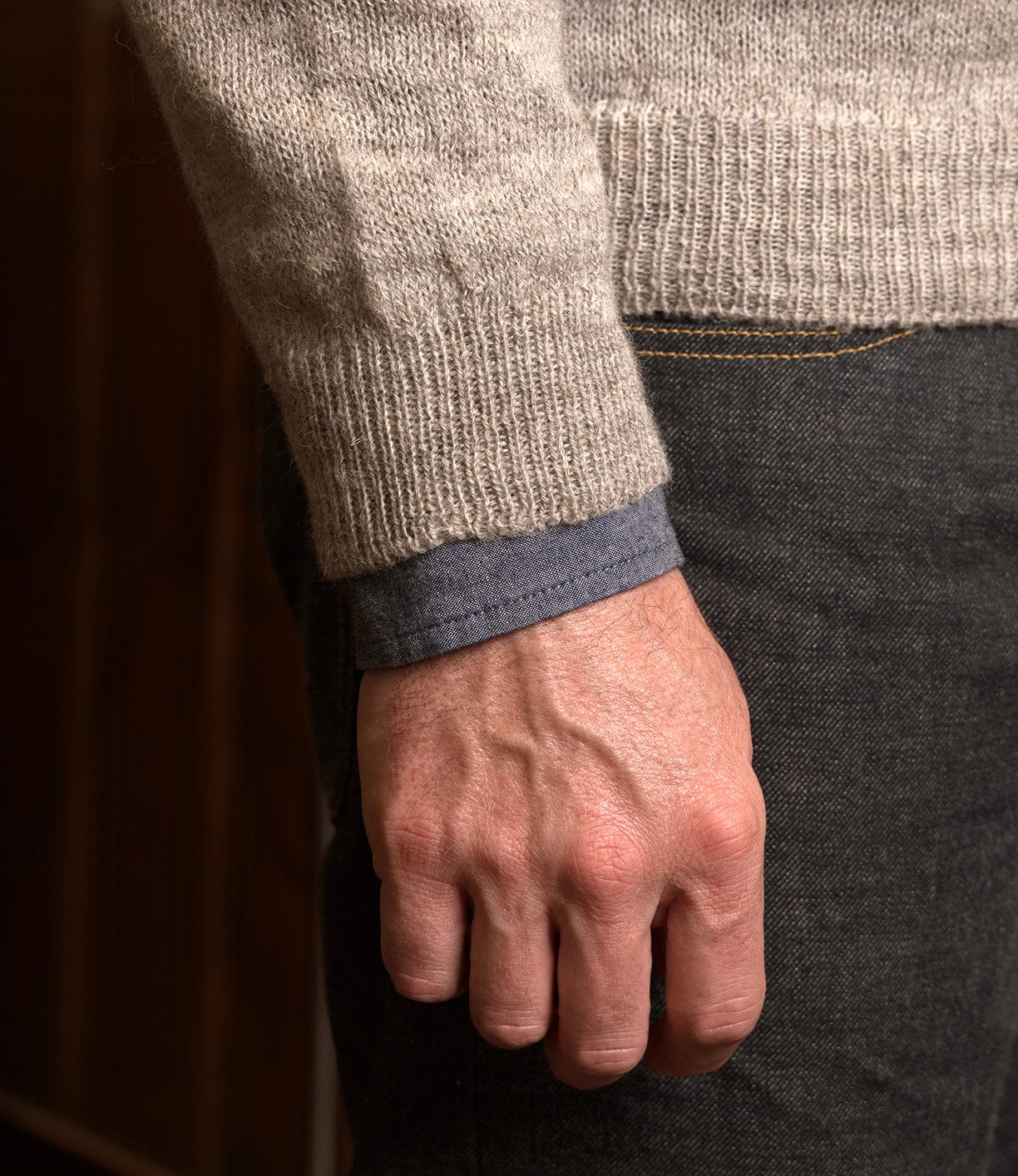 A close-up shows a person’s right hand resting by their side, wearing the Best Made Co. 5-Gauge Shetland Sweater in beige over a blue shirt and dark gray pants, with the softly blurred background highlighting the sweater’s lightweight warmth.