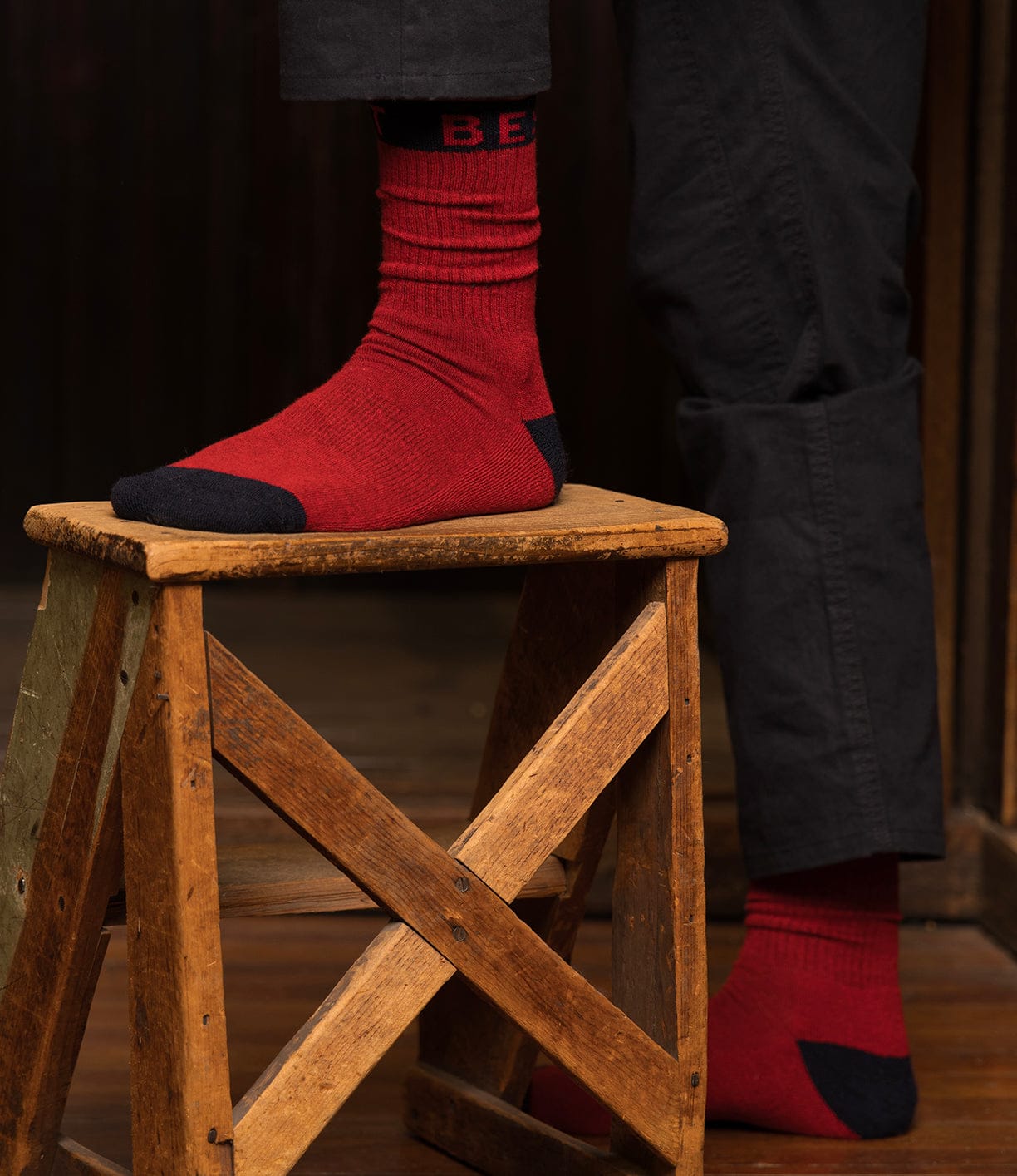 Wearing Best Made Co Standard Wool Socks featuring red bodies with dark toes and heels, a person in dark pants stands with one foot on a wooden stool, enjoying all-season comfort against dim wooden walls and floor.