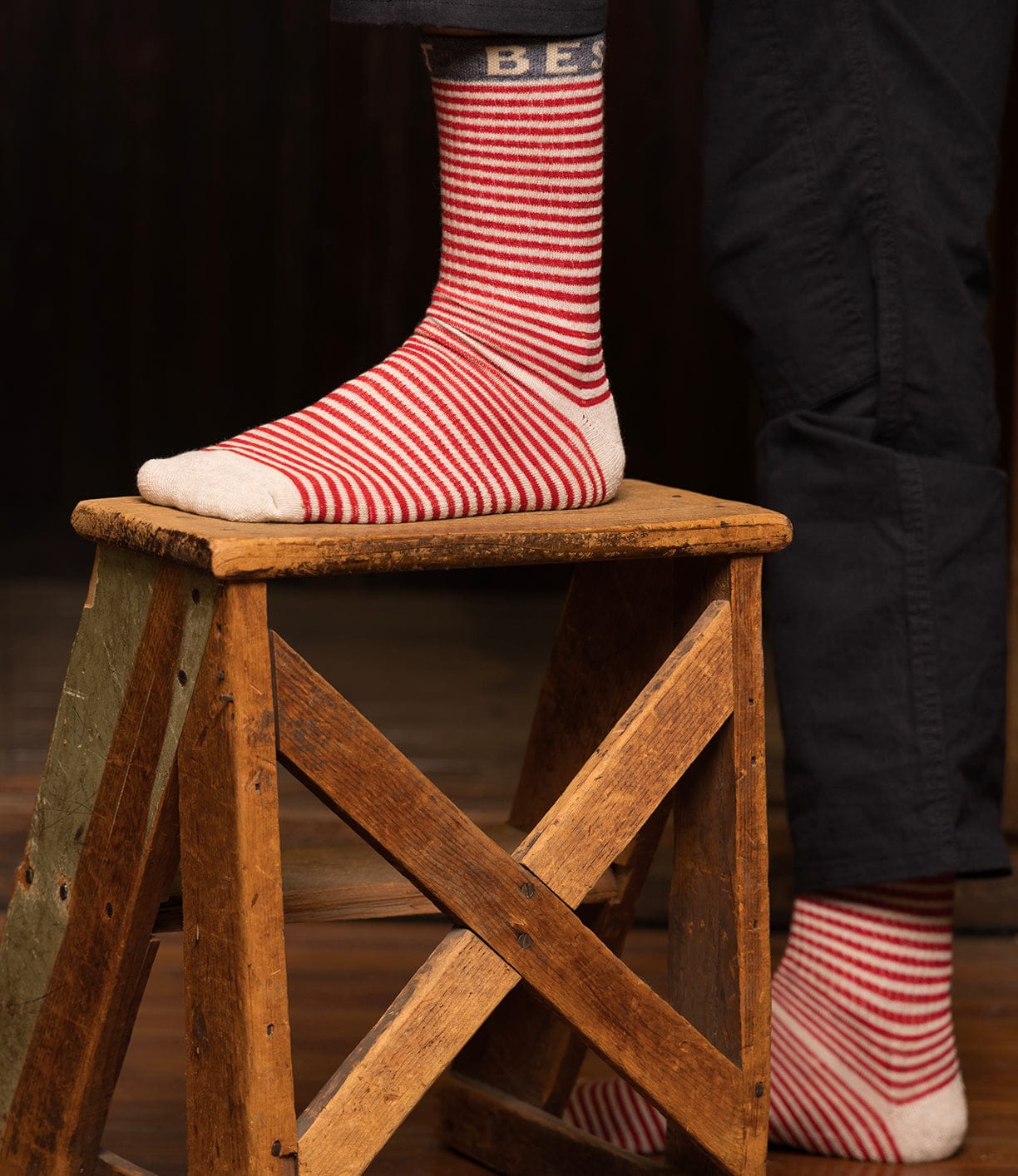 A person wearing Best Made Co Standard Wool Socks—red and white striped with “BEST” near the top—stands with one foot on a wooden step stool. These Merino wool socks provide premium moisture wicking and all-season comfort.